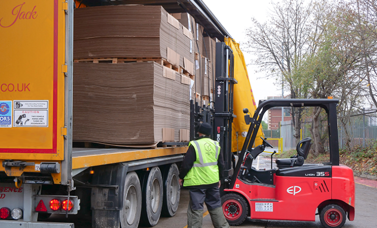 Red forklift truck with operator stood next to it loading flatpack corrugated cardboard boxes onto yellow lorry