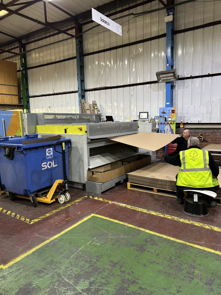 Packaging machine with two operators handling flat cardboard boxes that are coming out of it.